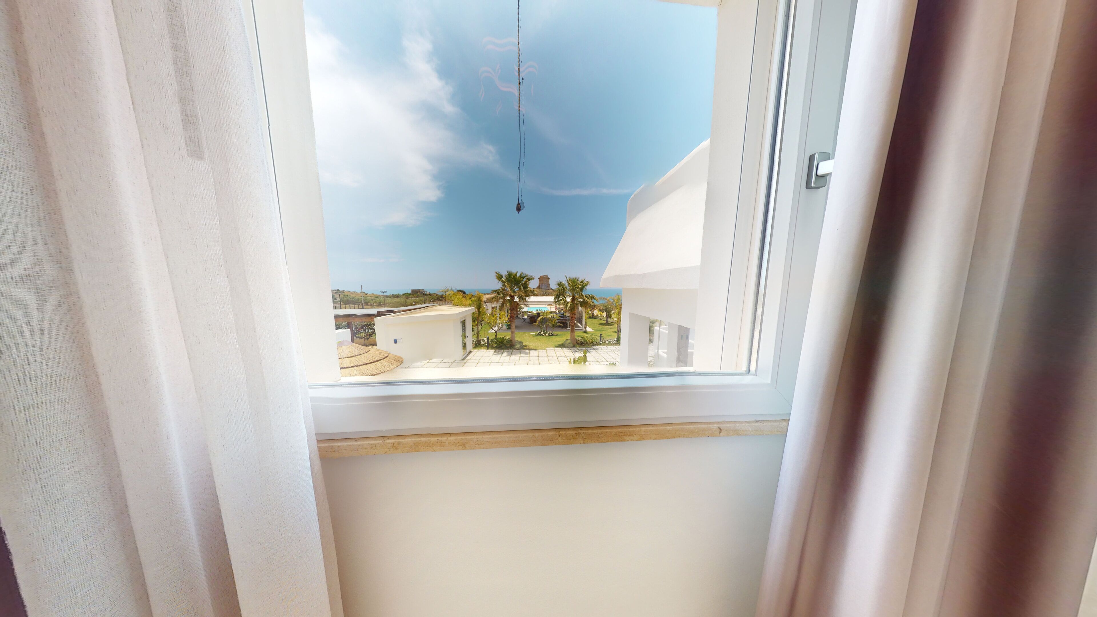 Une vue depuis une fenêtre à cadre blanc et rebord en bois, donnant sur une cour intérieure avec des palmiers, un bâtiment blanc et un ciel bleu dégagé.