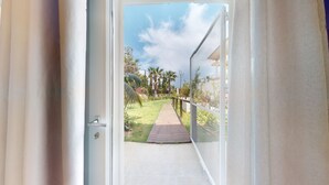 On aperçoit, à travers une porte ouverte, un chemin pavé qui mène à une zone herbeuse bordée de palmiers, avec un bâtiment en arrière-plan.