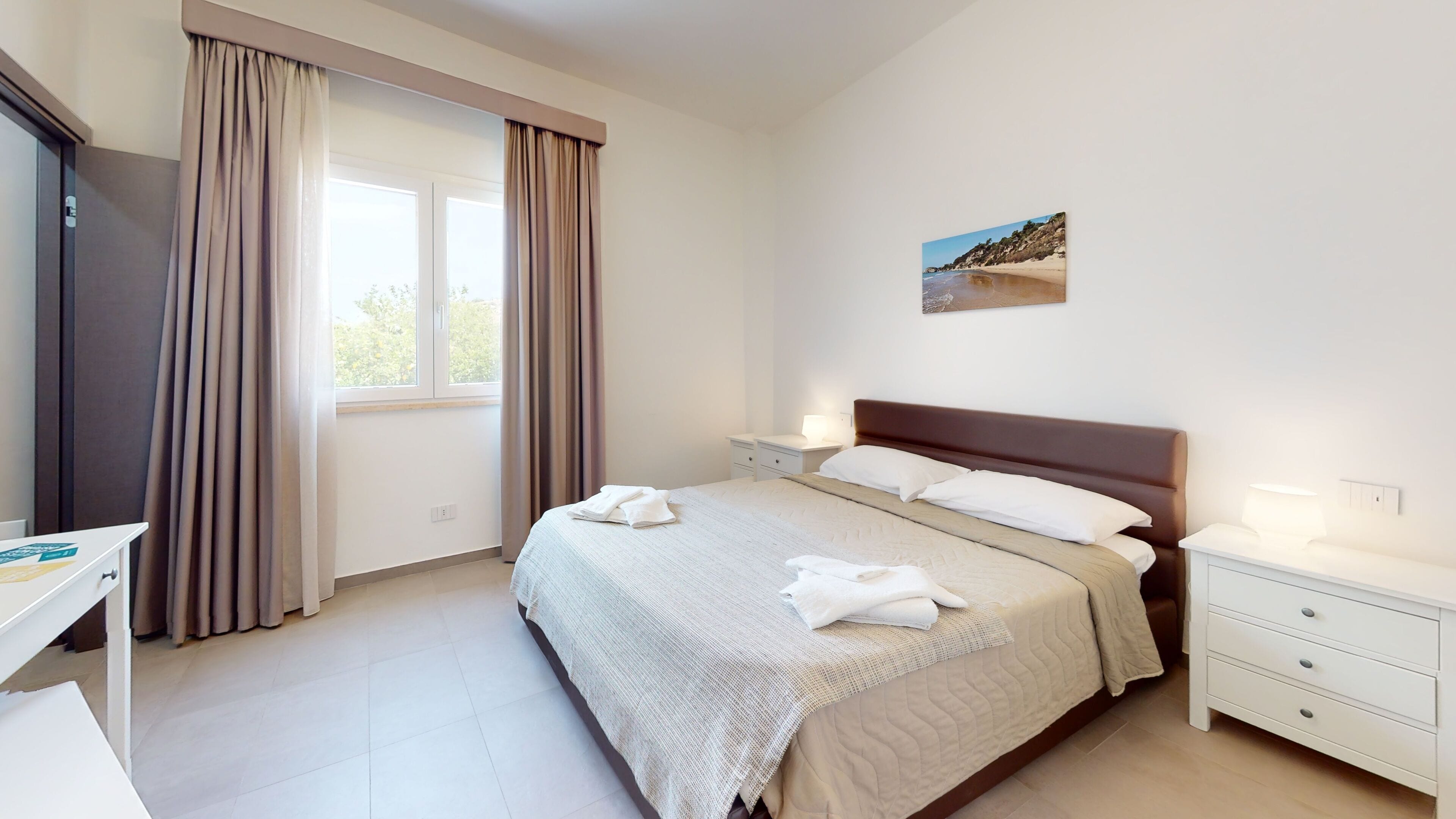 A hotel room with a bed, bedside tables, a desk, and a window with curtains.