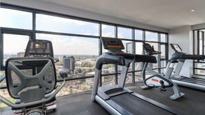 Ett gym med löpband, en crosstrainer och en motionscykel, med utsikt över staden genom stora fönster.