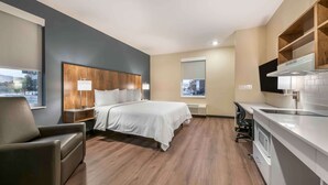 Une chambre d’hôtel moderne avec un lit doté d’une tête de lit en bois, un bureau avec une chaise, une grande fenêtre et un coin cuisine.