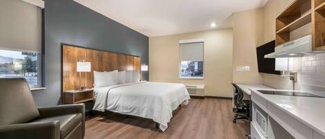 A modern hotel room with a wooden headboard bed, a desk with a chair, a large window, and a kitchenette.