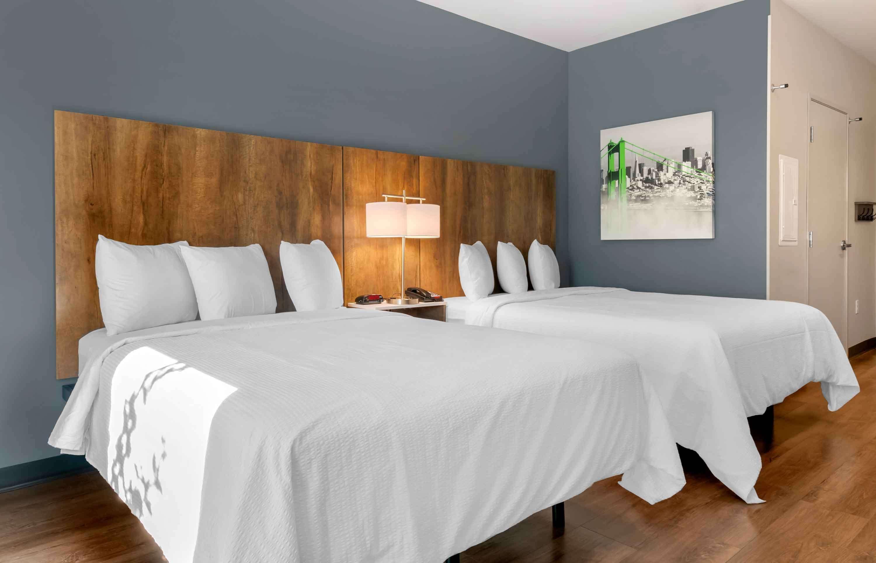 Une chambre d’hôtel avec deux lits, une tête de lit en bois, une table de chevet avec une lampe et un tableau représentant un paysage urbain accroché au mur.
