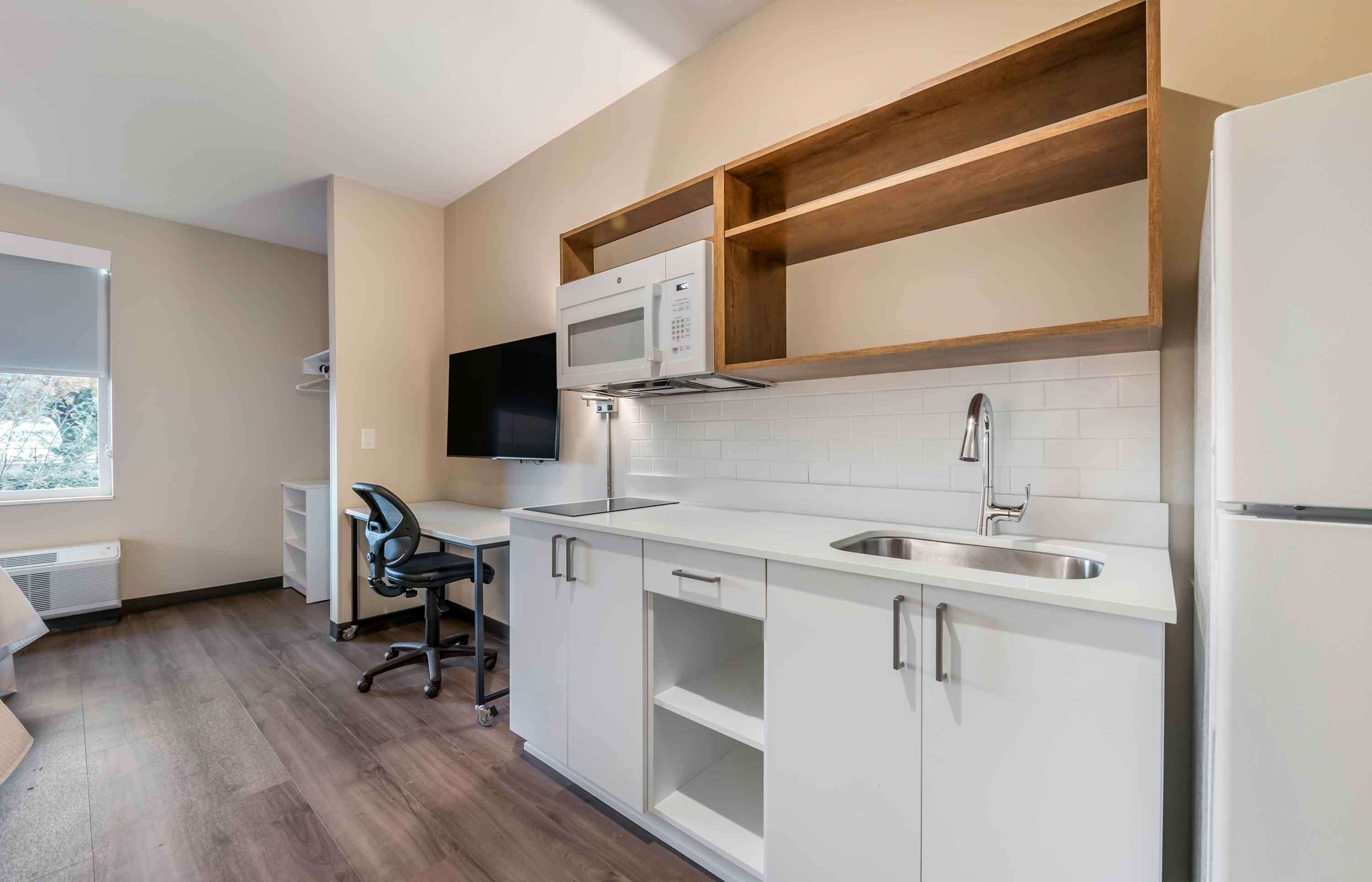 A modern kitchen with white cabinets, a microwave, and a built-in oven. There is a window providing natural light and a chair in the corner.