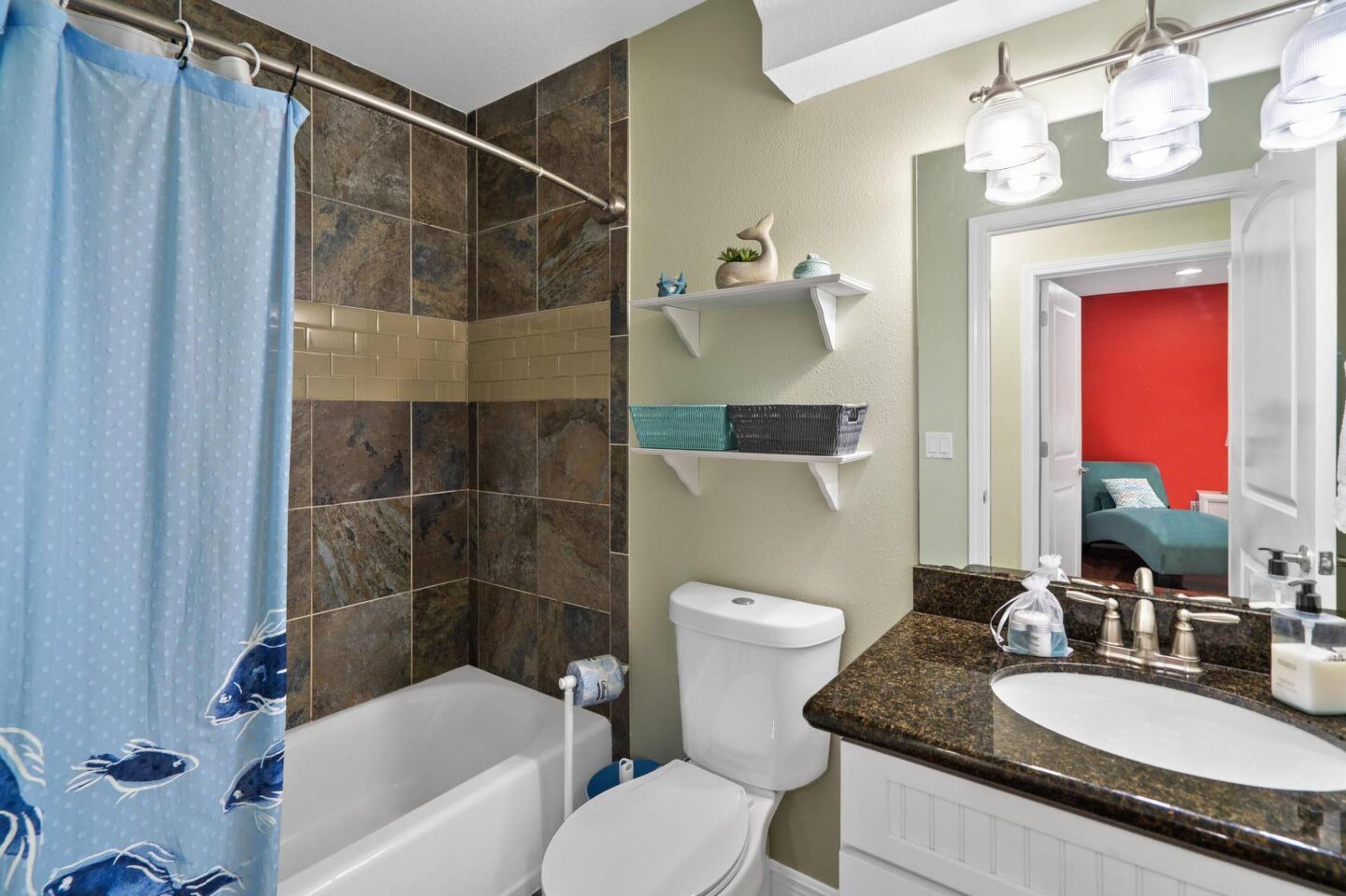 A bathroom with a shower, toilet, sink, and a mirror with lights. There are shelves with toiletries and a red wall in the background.