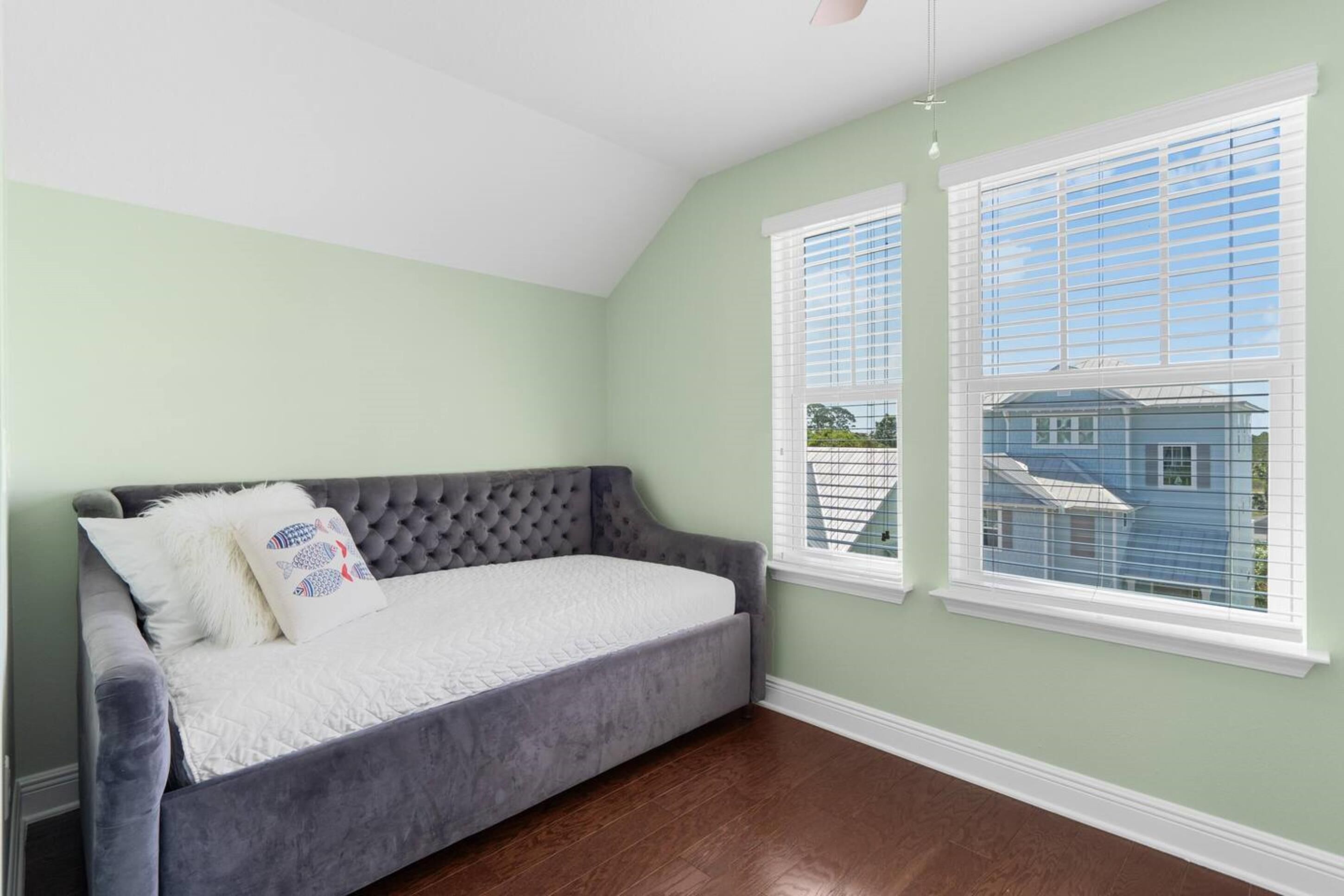 A room with a grey sofa bed, two windows with white blinds, and a view of a blue house outside.