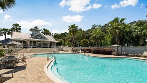 A swimming pool with lounge chairs and a covered seating area, surrounded by palm trees and a building with a distinctive roof. - 30A Beach House - Pelican's Rest by PHG (Panama City Beach)