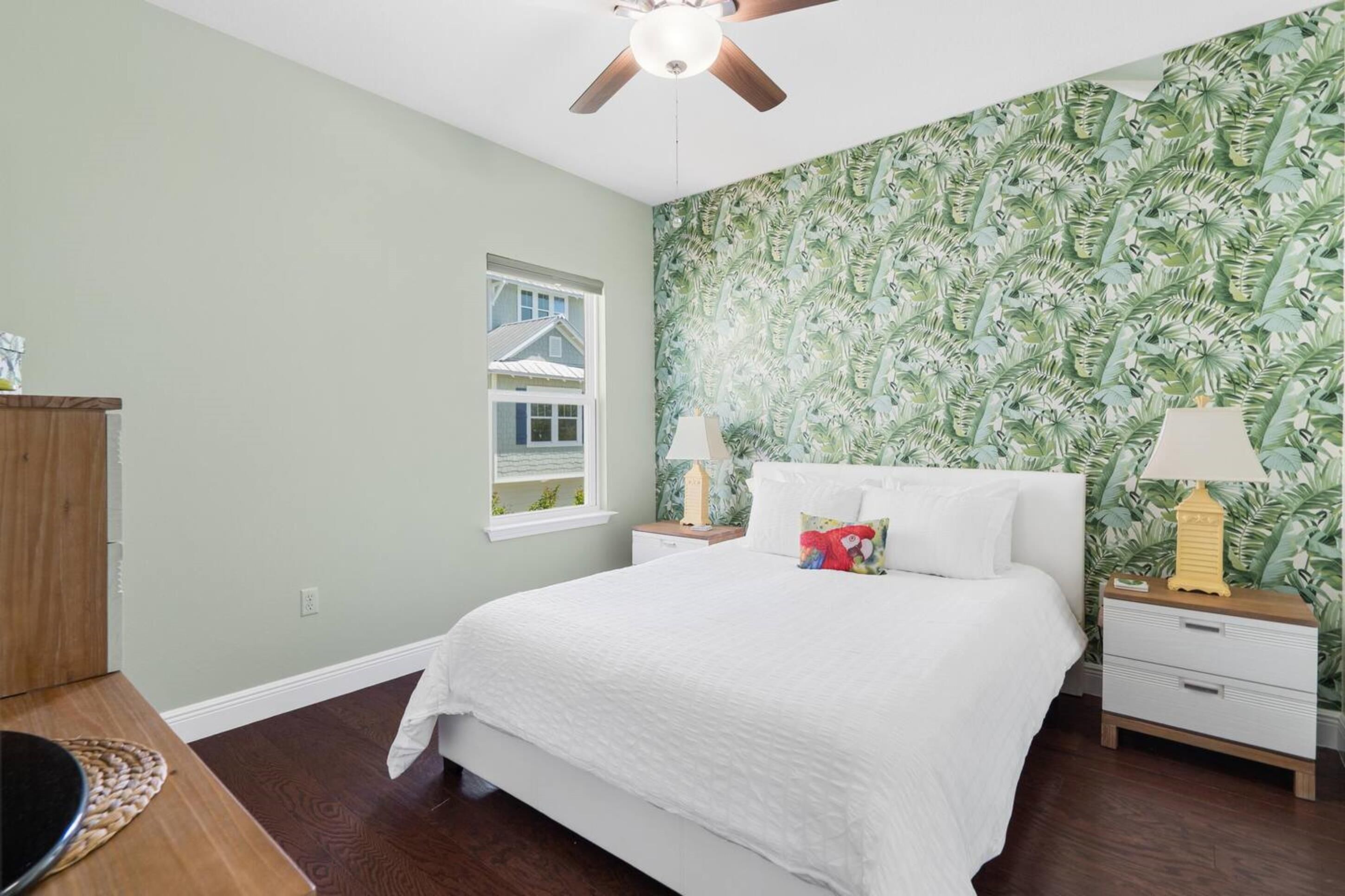 A bedroom with a large bed, two bedside tables with lamps, a ceiling fan, a window with a view of a house, and a wall with a leafy pattern.
