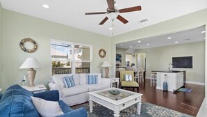 A living room with a white sofa, blue armchair, and a coffee table. There is a ceiling fan, a window with blinds, and a kitchen area in the background. - 30A Beach House - Pelican's Rest by PHG (Panama City Beach)
