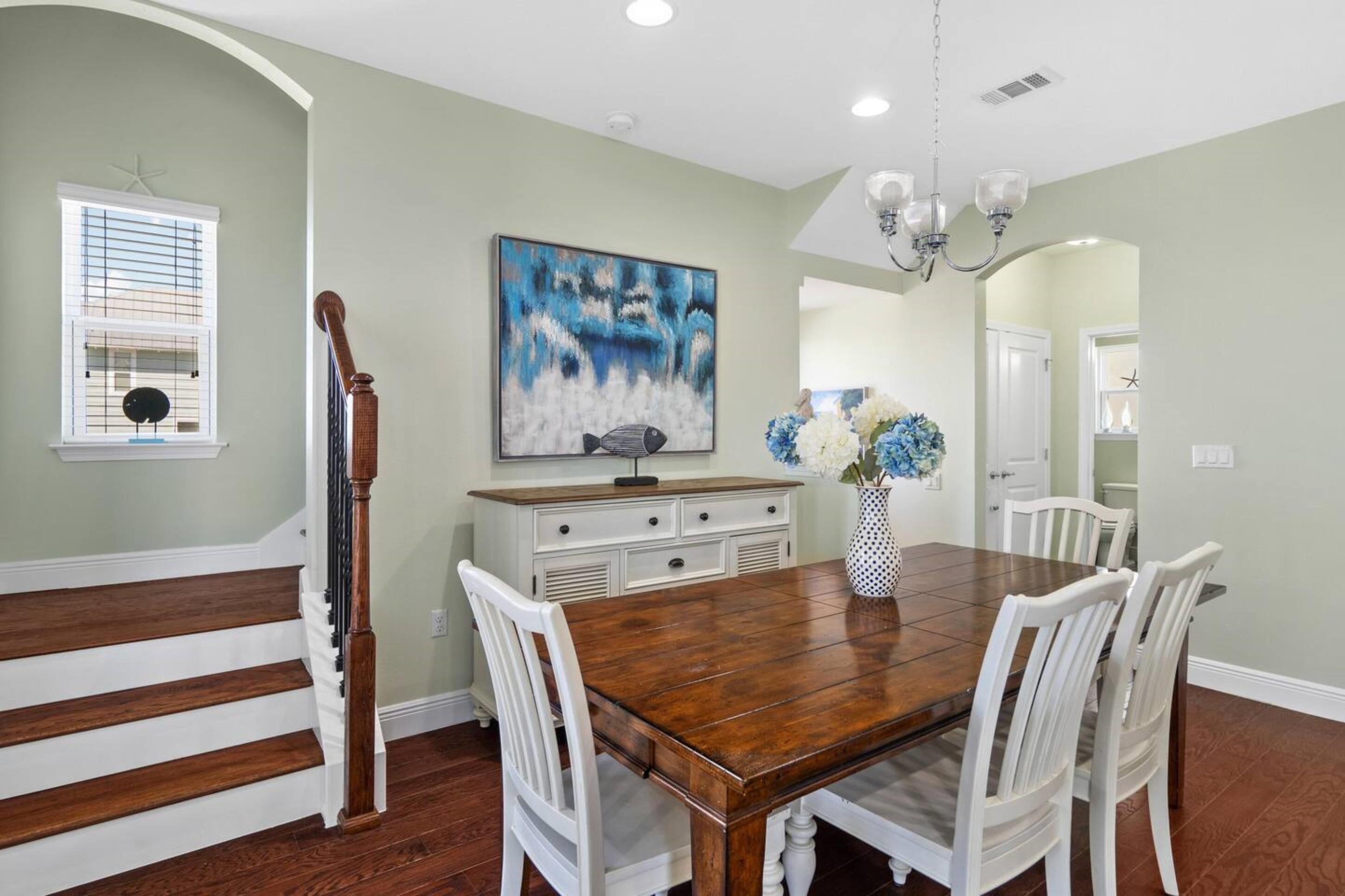 Uma área de refeições com mesa de madeira e cadeiras brancas, um armário branco com gavetas, um quadro na parede e um candelabro.