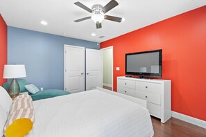 Une chambre à coucher avec un lit, un ventilateur de plafond, un téléviseur posé sur une commode blanche et une porte donnant sur un placard.