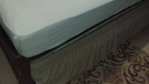 Uma cama com colchão e almofadas claras, cabeceira de madeira e tapete estampado no chão.