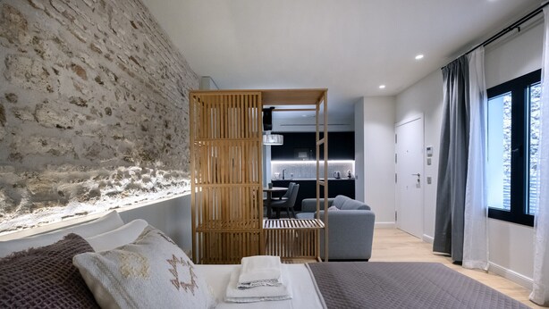 Een moderne hotelkamer met een stenen muur, een houten scheiding, een bed met grijze deken en uitzicht op de keuken en eetruimte.