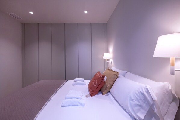 A hotel room with a neatly made bed, a wall-mounted lamp, and a ceiling light.