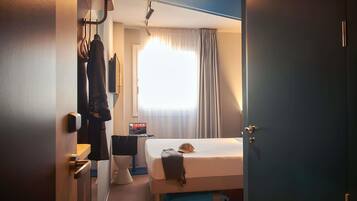 Et hotelværelse med seng, kuffert, skrivebord og et vindue med gardiner.
