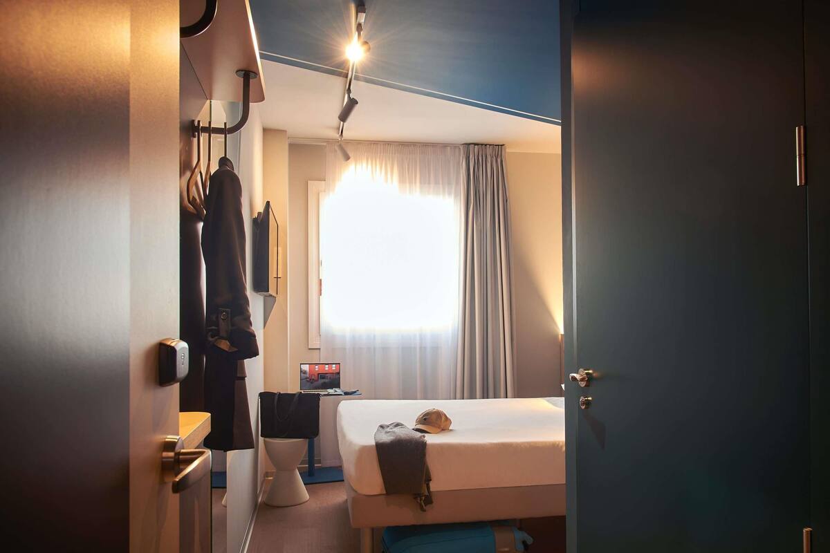 Una habitación de hotel con cama, maleta, escritorio y ventana con cortinas.