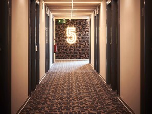 Un couloir avec un tapis à motifs, un mur orné d’un motif géométrique et un panneau lumineux représentant le chiffre « 5 ».
