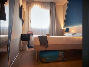 A hotel room with a bed, a desk with a computer, a suitcase, and a window with curtains.