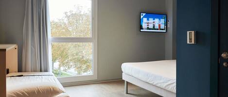 Una habitación con una cama, un televisor montado en la pared y un ventanal con cortinas.