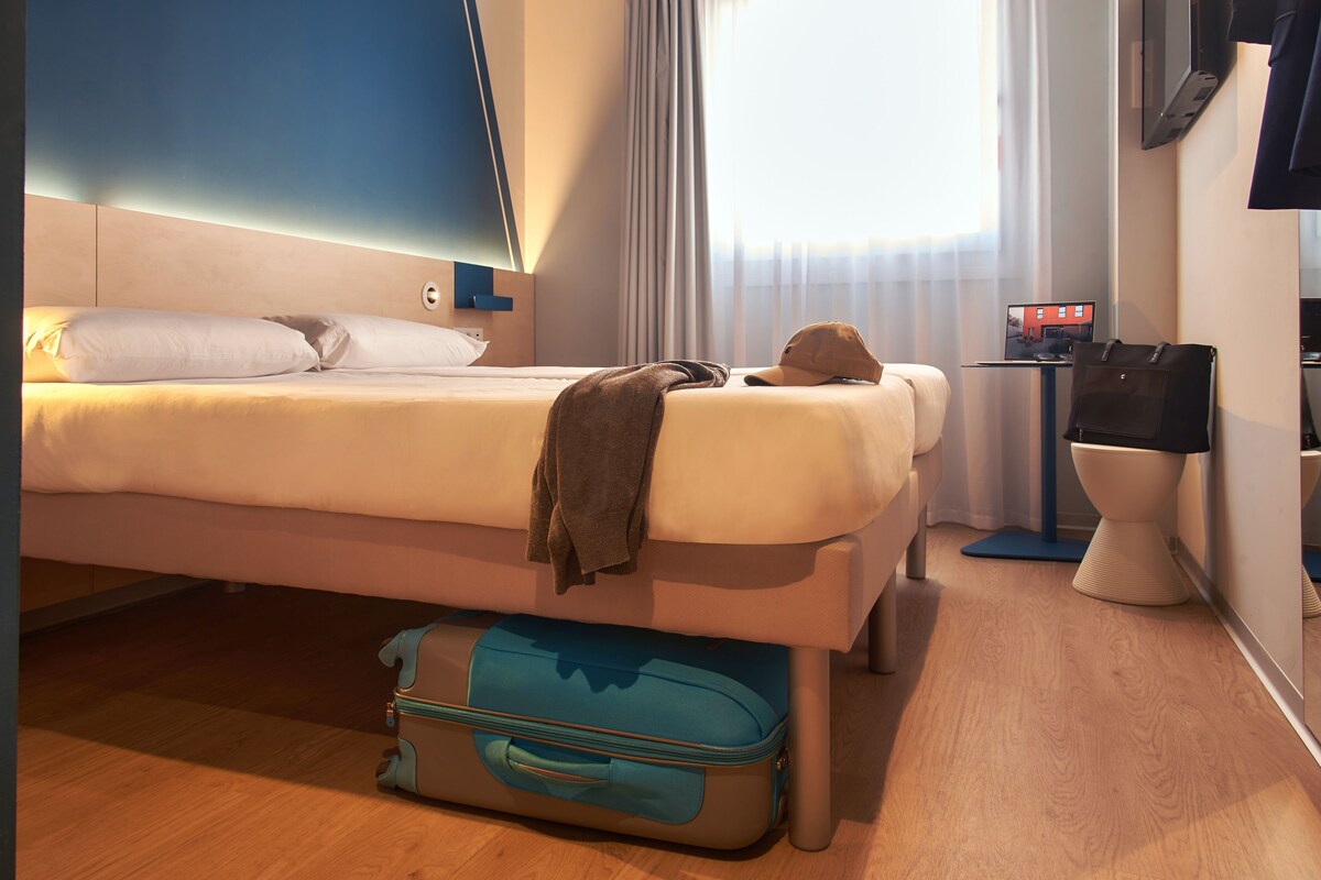 Une chambre d’hôtel avec un lit, une valise, un bureau avec un ordinateur et une fenêtre avec des rideaux.