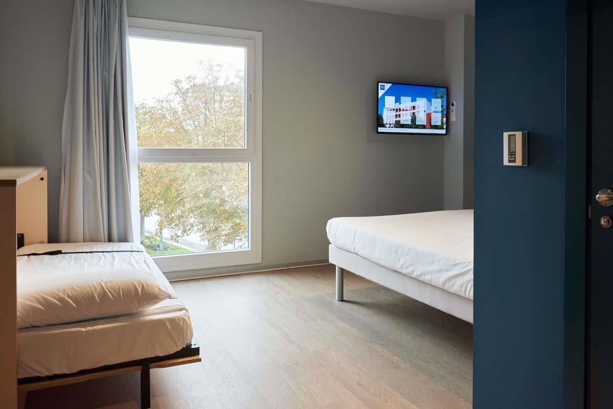 Una habitación con una cama, un televisor montado en la pared y un ventanal con cortinas.