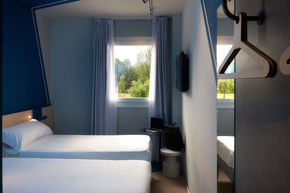 Une chambre d’hôtel avec un lit, un bureau, une chaise, une télévision et une fenêtre avec des rideaux.