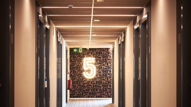 Un pasillo con piso estampado, el número '5' iluminado en la pared y puertas a ambos lados.