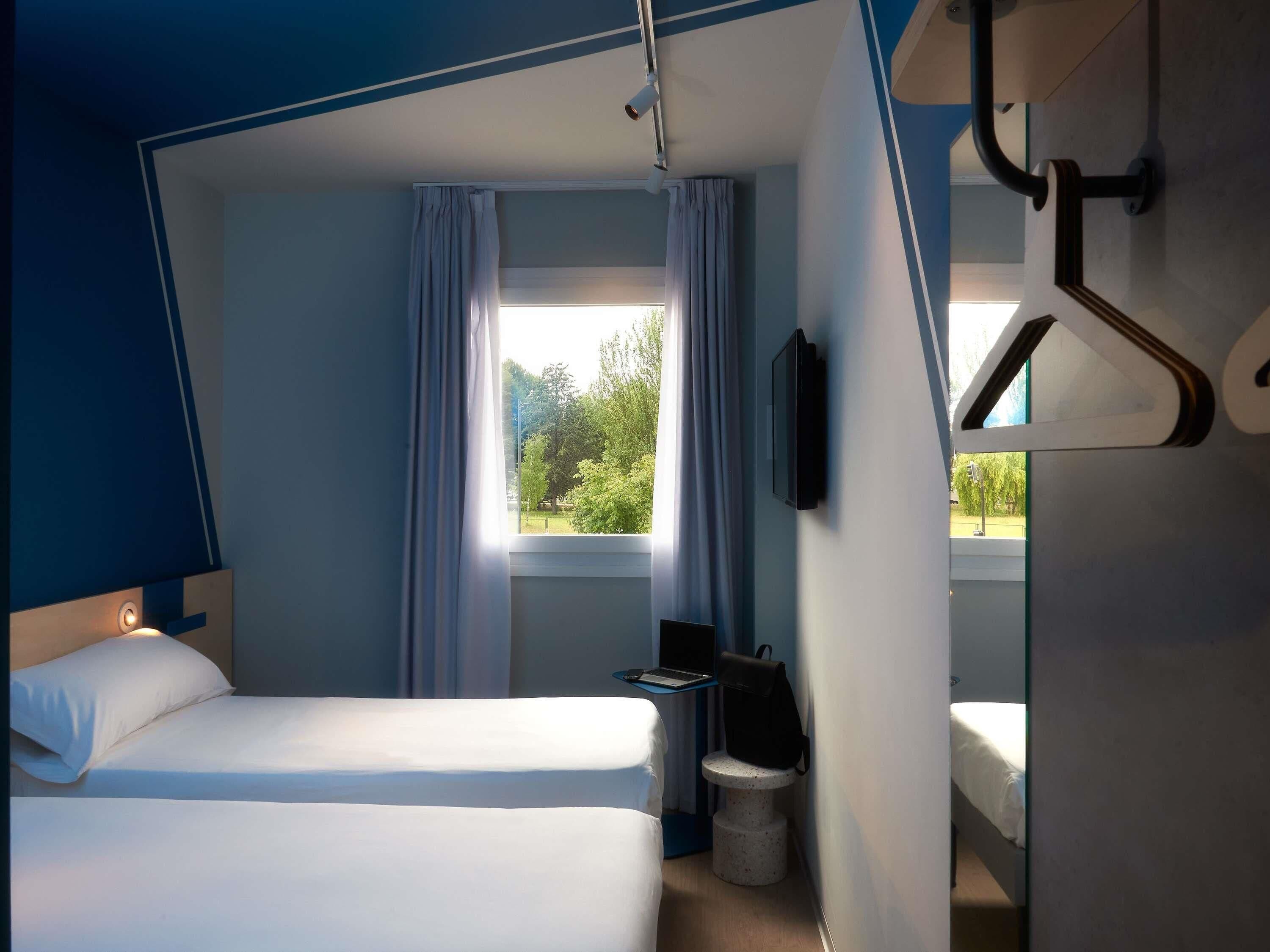 Una habitación de hotel con cama, escritorio, silla, televisor y ventana con cortinas.