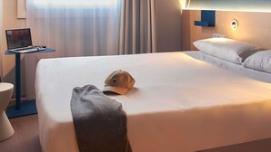 Habitación de hotel con una cama, una laptop sobre una mesita auxiliar, una toalla y un sombrero en la cama.