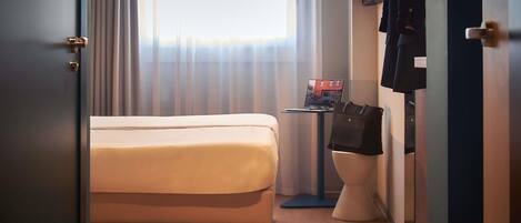 Una habitación de hotel con cama, una maleta, un ordenador portátil sobre una mesita y un inodoro.