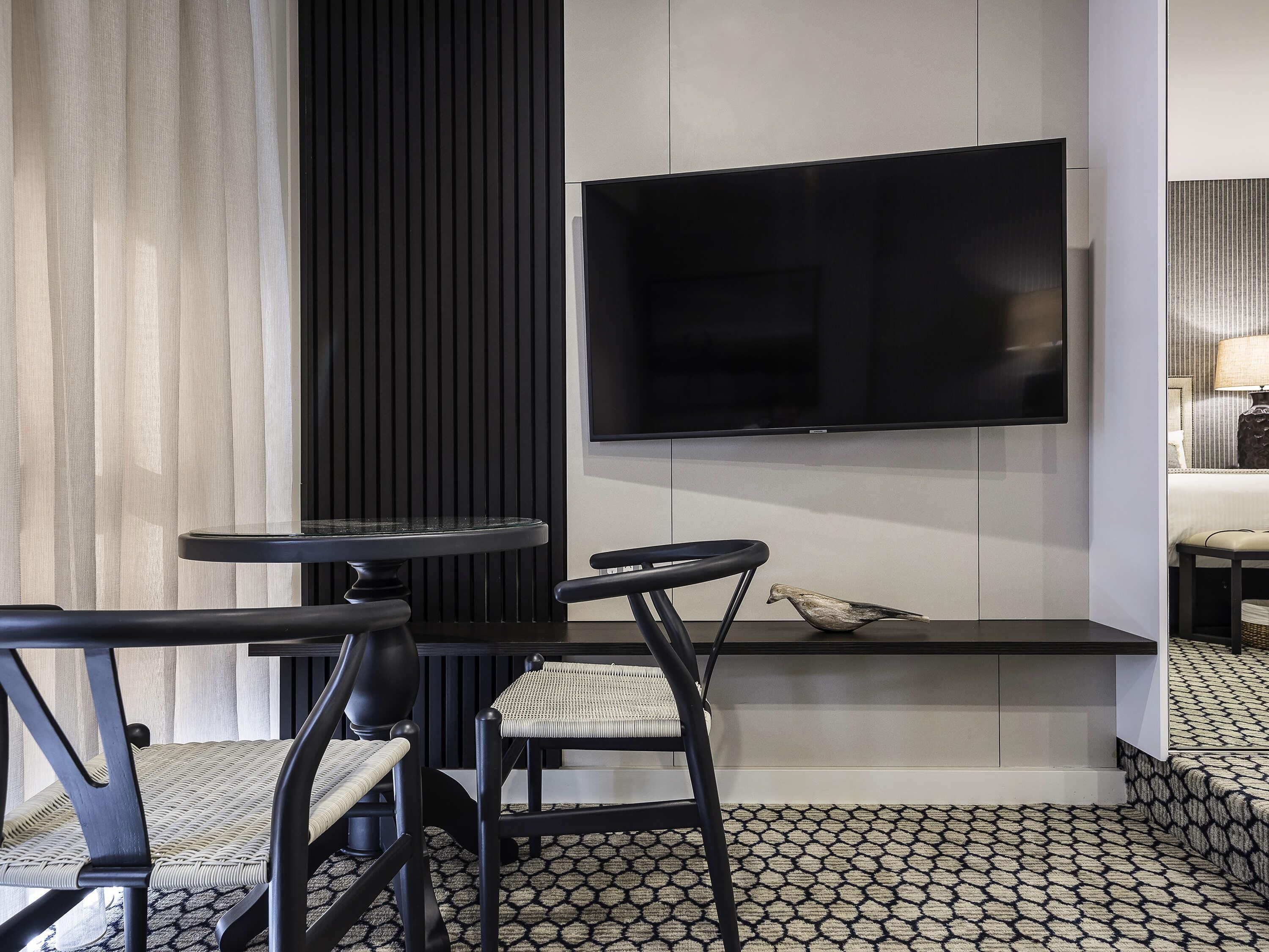 Una habitación de hotel moderna con un televisor de pantalla plana montado en la pared, una mesa de comedor redonda, dos sillas negras y un suelo estampado.