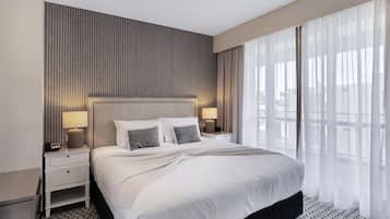 Una habitación de hotel moderna con una cama grande, dos mesitas de noche con lámparas, un cabecero y vistas a través de cortinas traslúcidas.
