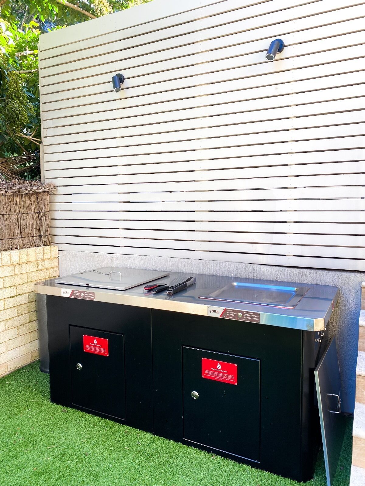 A barbecue grill with a black cabinet and a metal top, placed outdoors with a white wall and green grass in the background.