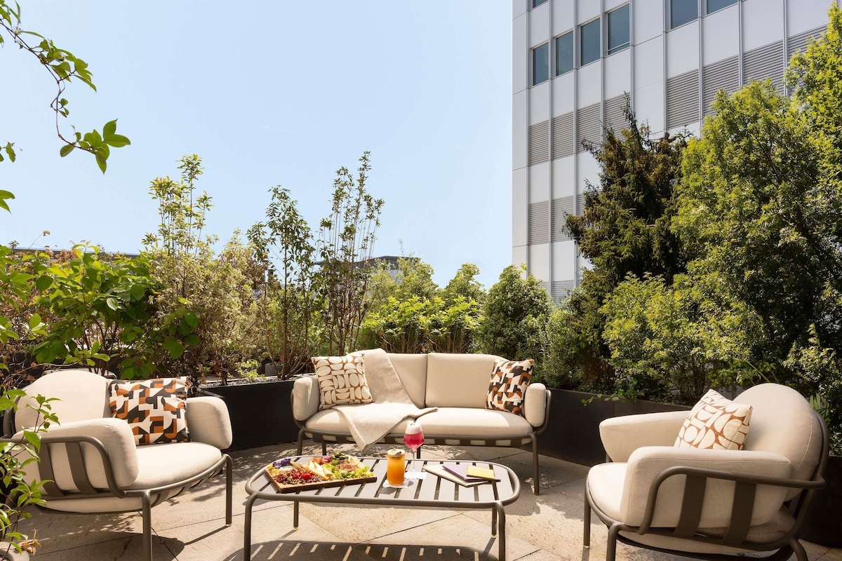 An outdoor seating area with a sofa set and a coffee table, surrounded by greenery and a modern building in the background.