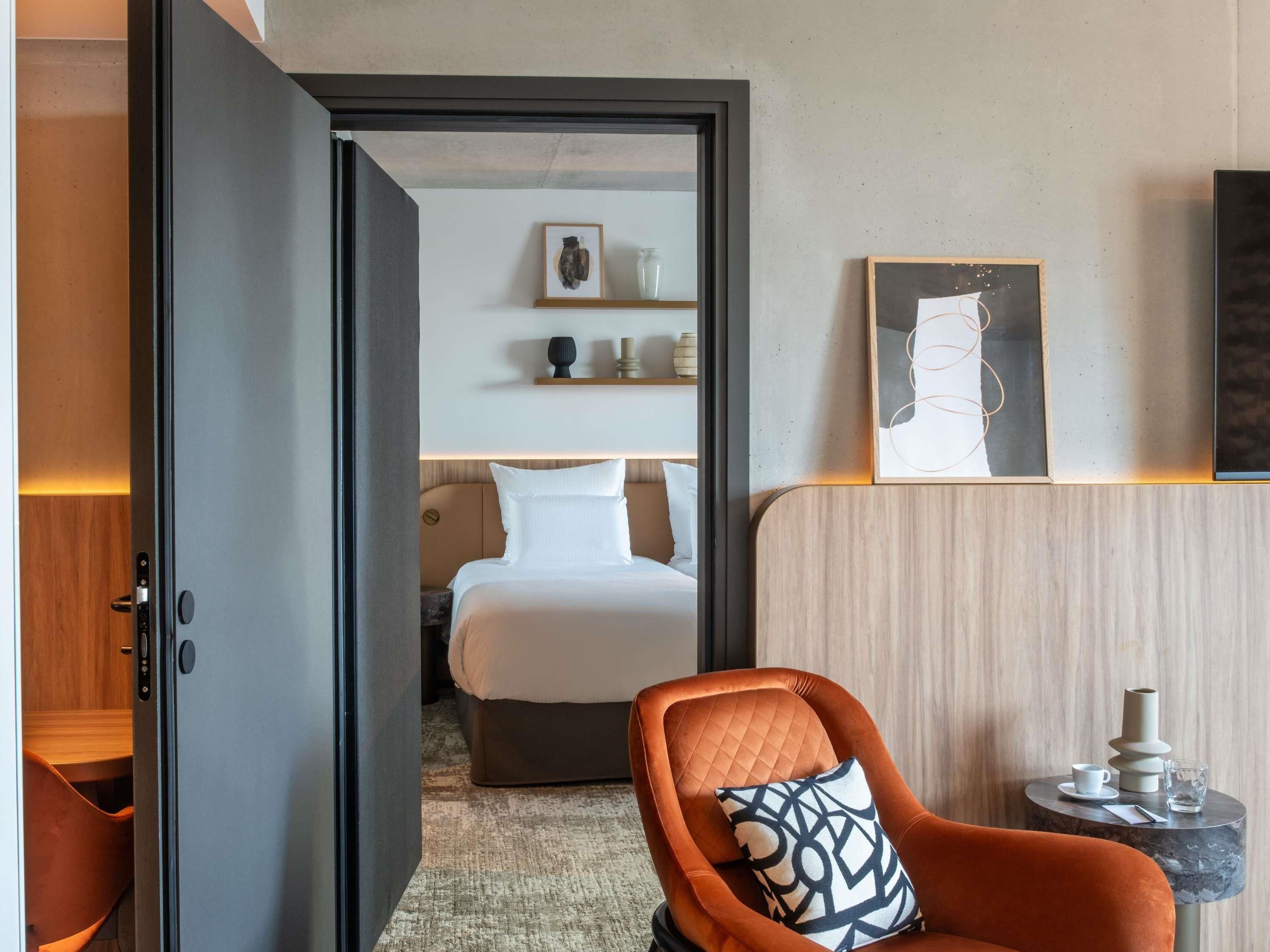 Una habitación de hotel con una cama, una silla con cojín estampado, una mesita con una taza y un jarrón, y una obra de arte en la pared.