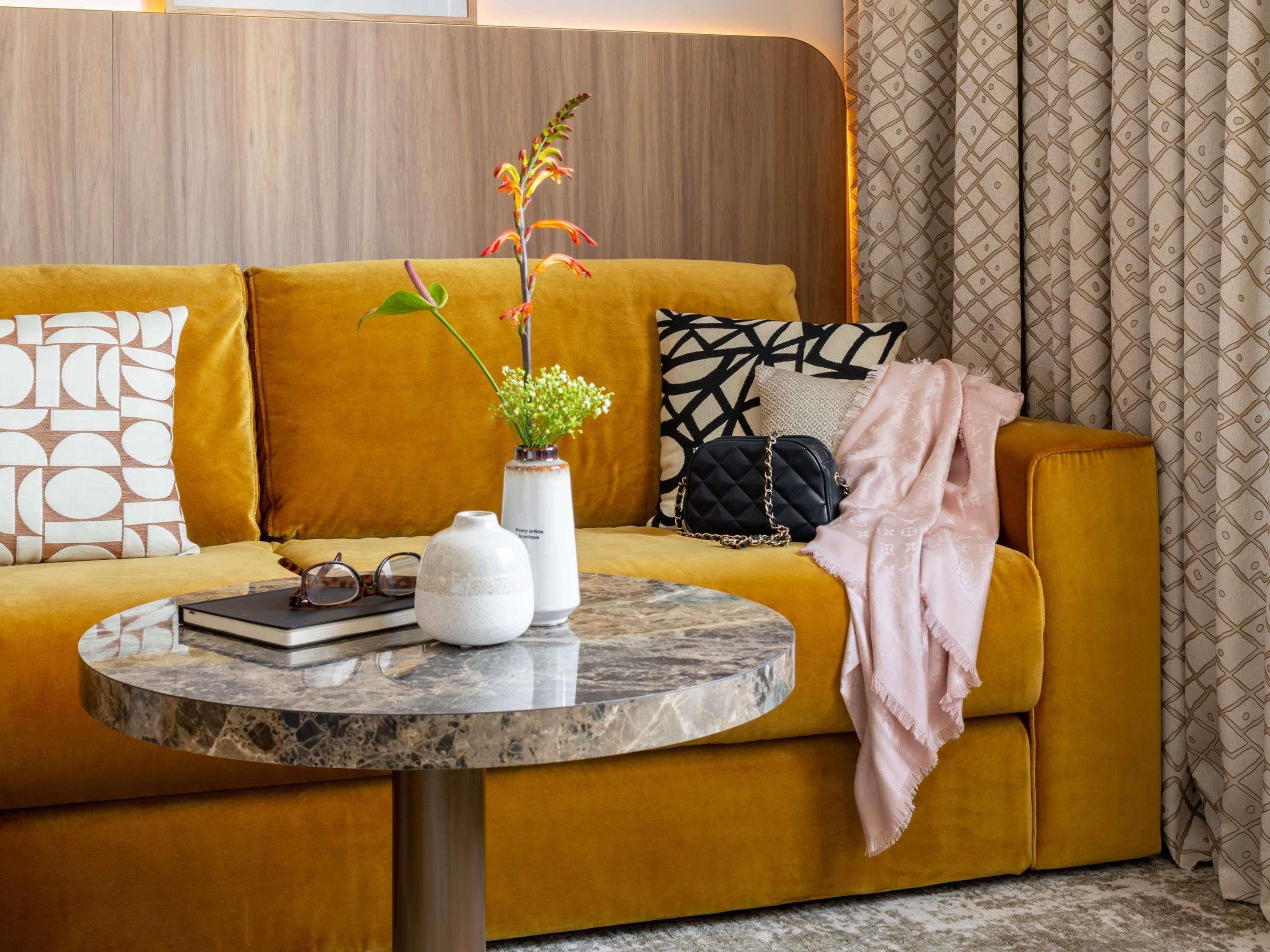 A mustard-colored sofa with a marble side table, a white vase with flowers, a black quilted bag, and patterned pillows.