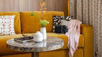 A mustard-colored sofa with a marble side table, a white vase with flowers, a black quilted bag, and patterned pillows.