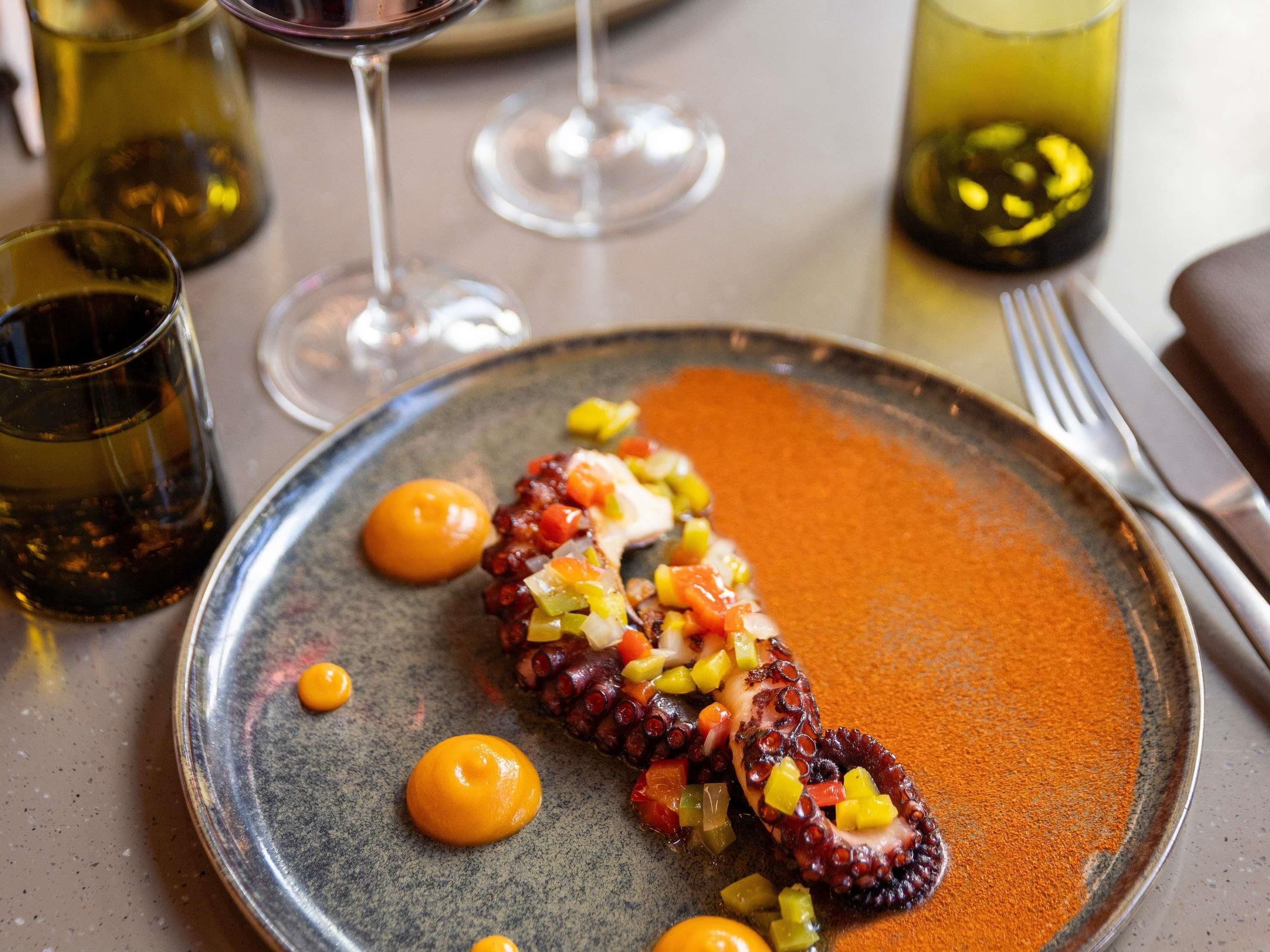 A plate with octopus and vegetables, a fork, and a glass of wine on a table.