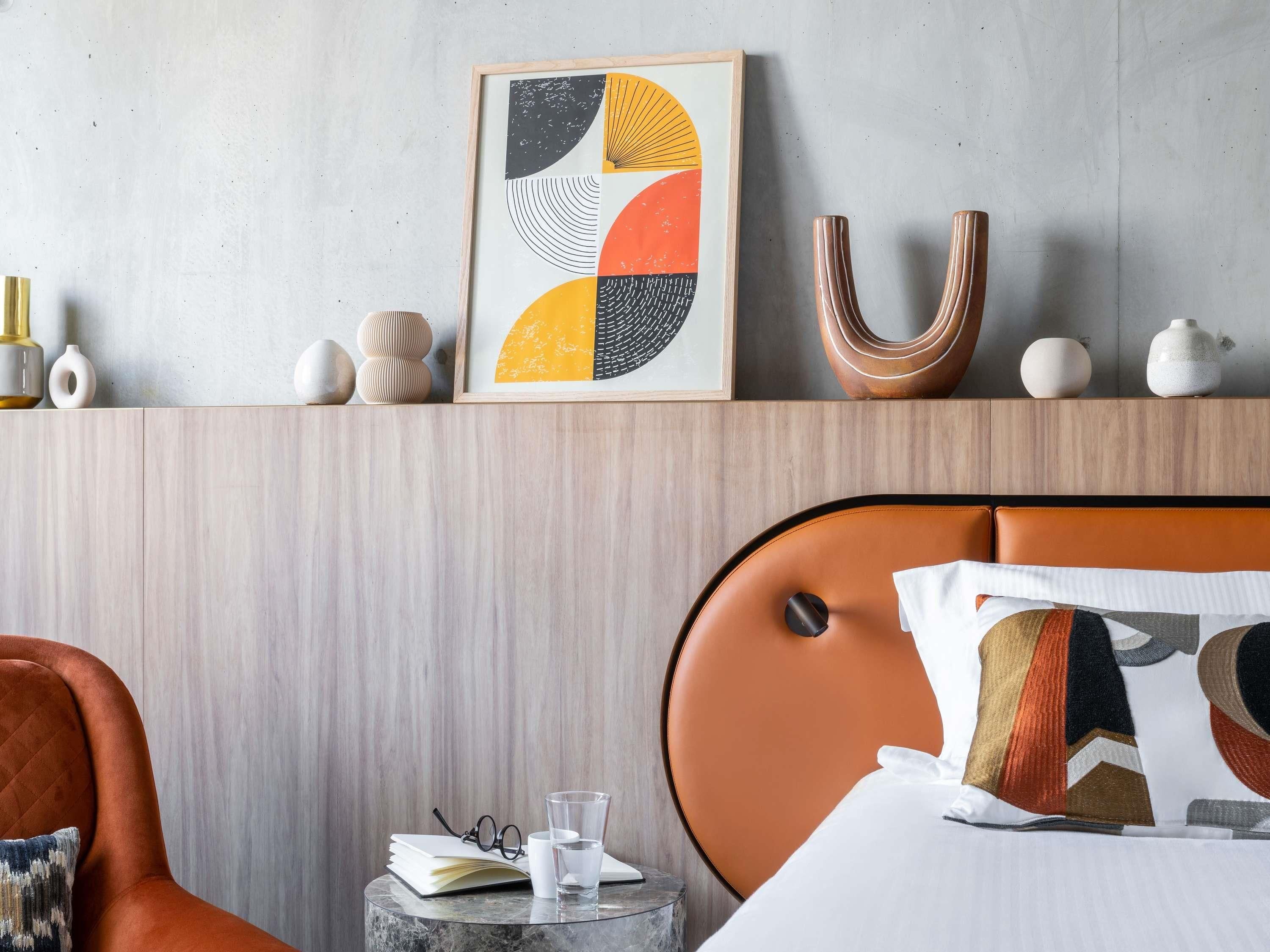 Una habitación de hotel moderna con cabecera de madera, una obra de arte abstracta enmarcada, una mesita de noche de mármol y un sillón de cuero.