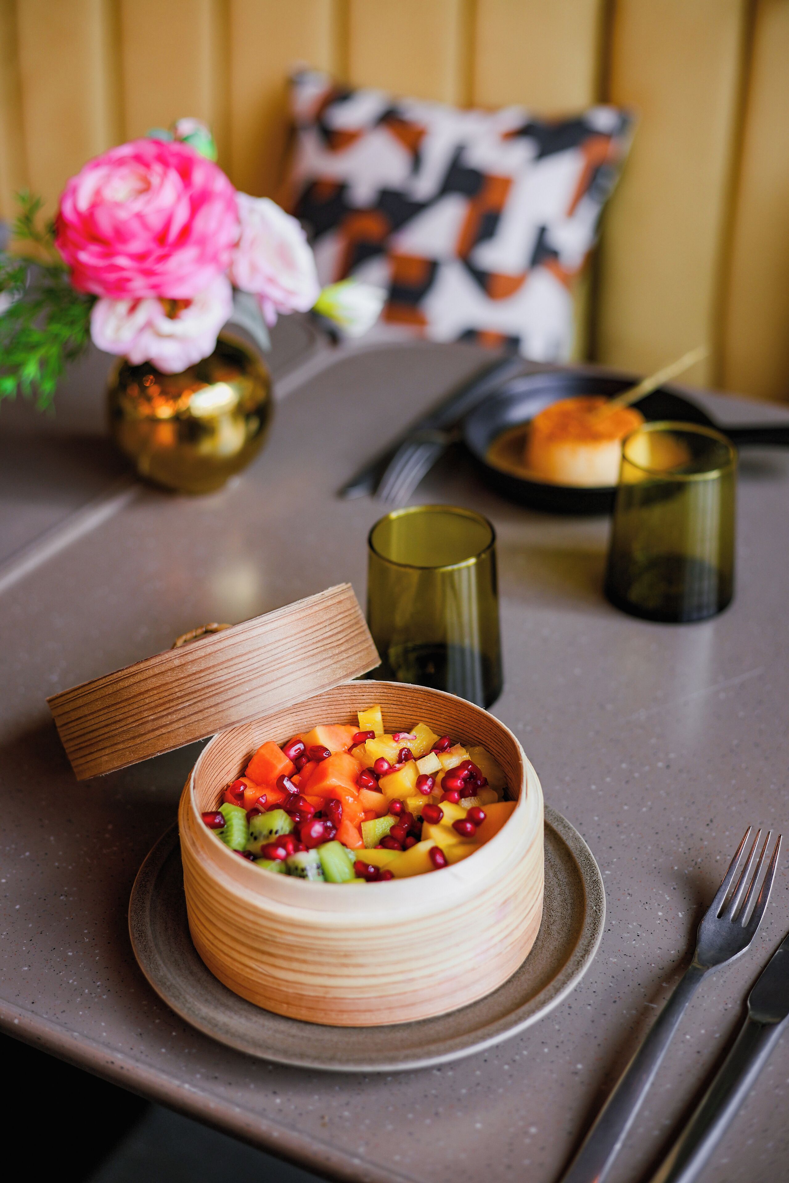 Een houten schaal met deksel gevuld met fruitsalade, geplaatst op een tafel met servet, vork en glas.