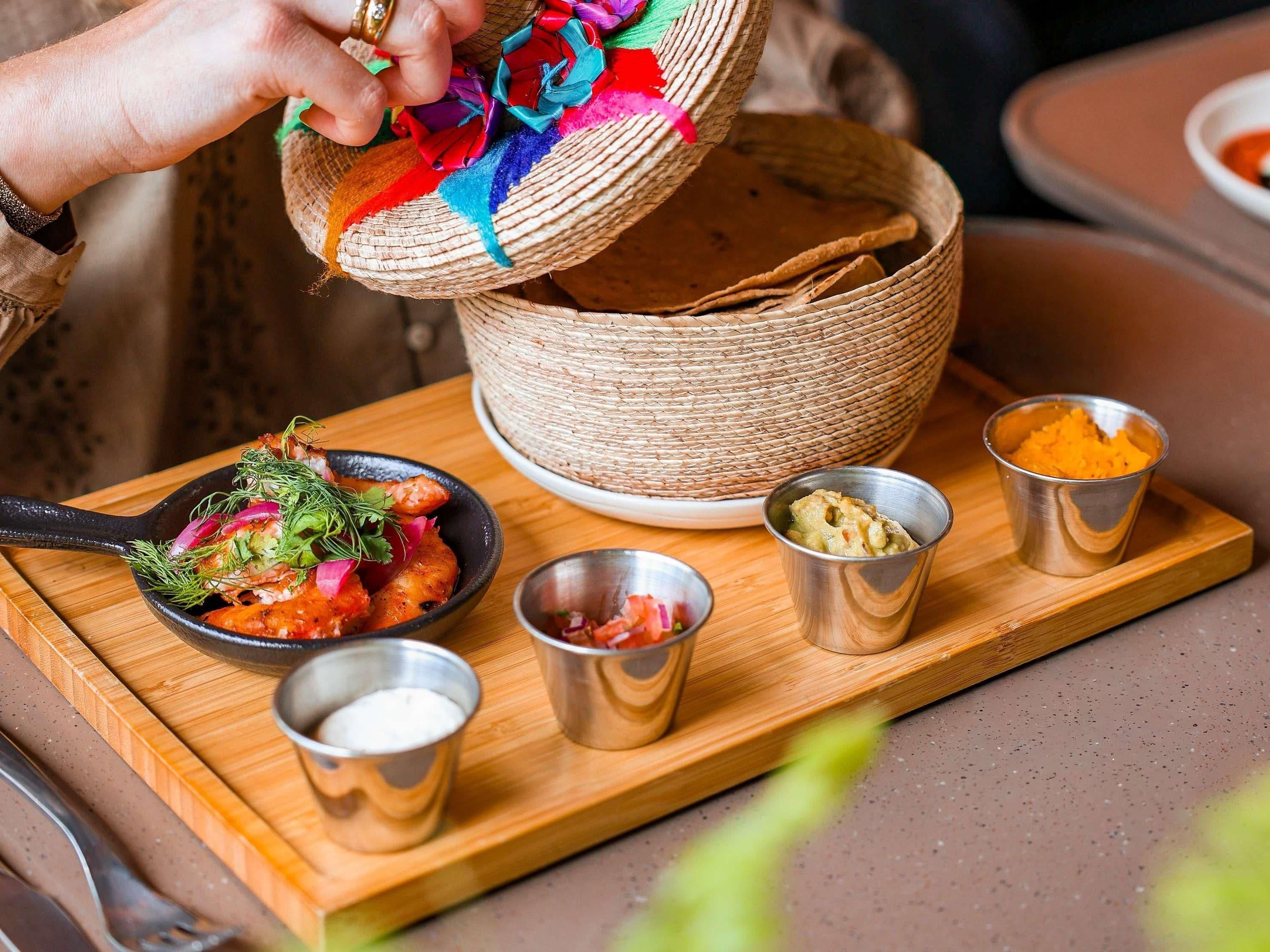 A person pouring colorful fabric into a woven basket on a wooden board with food items and small metal cups.
