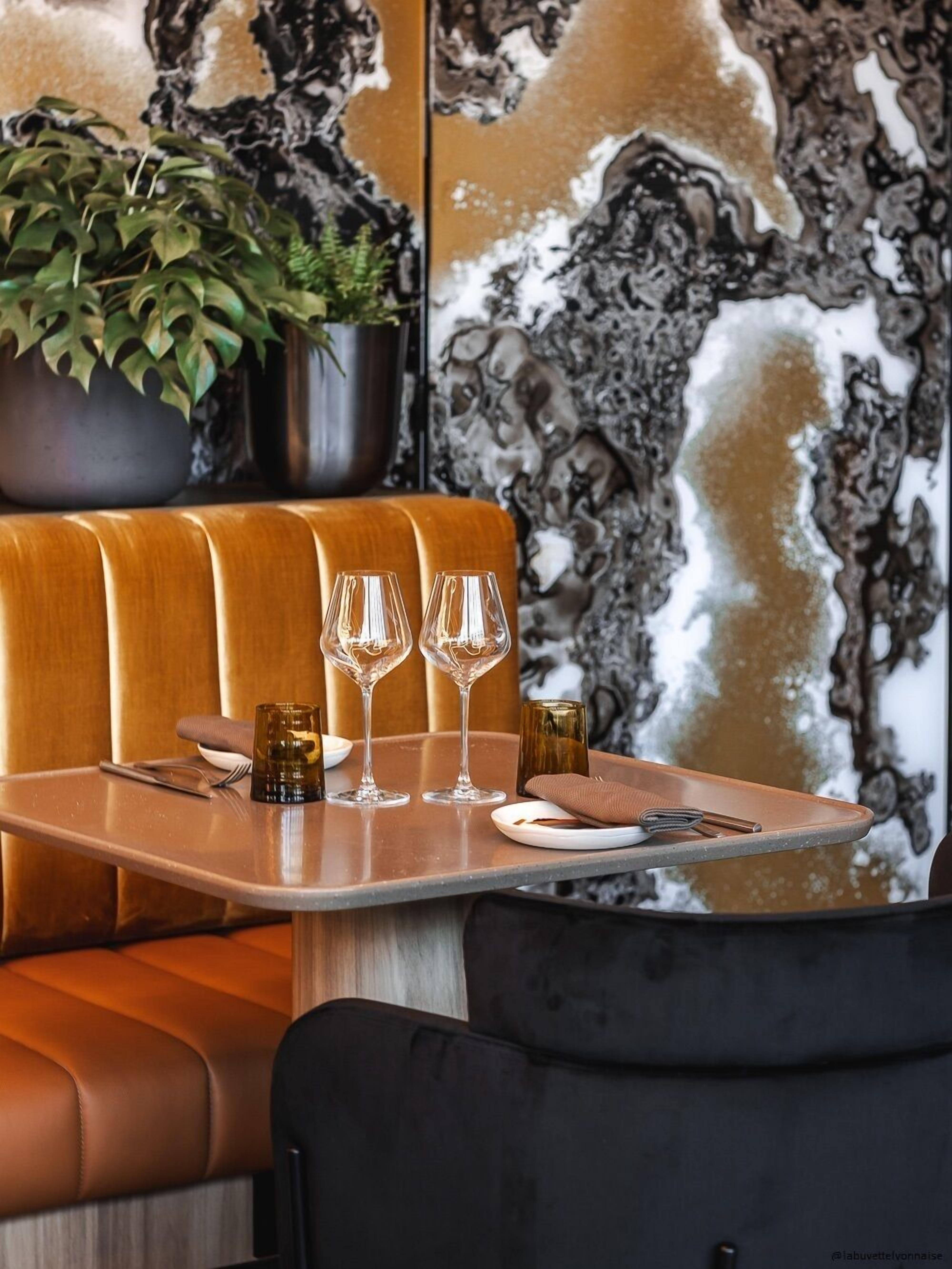 A restaurant booth with a marble wall backdrop, a potted plant, and a table set with wine glasses and a menu.