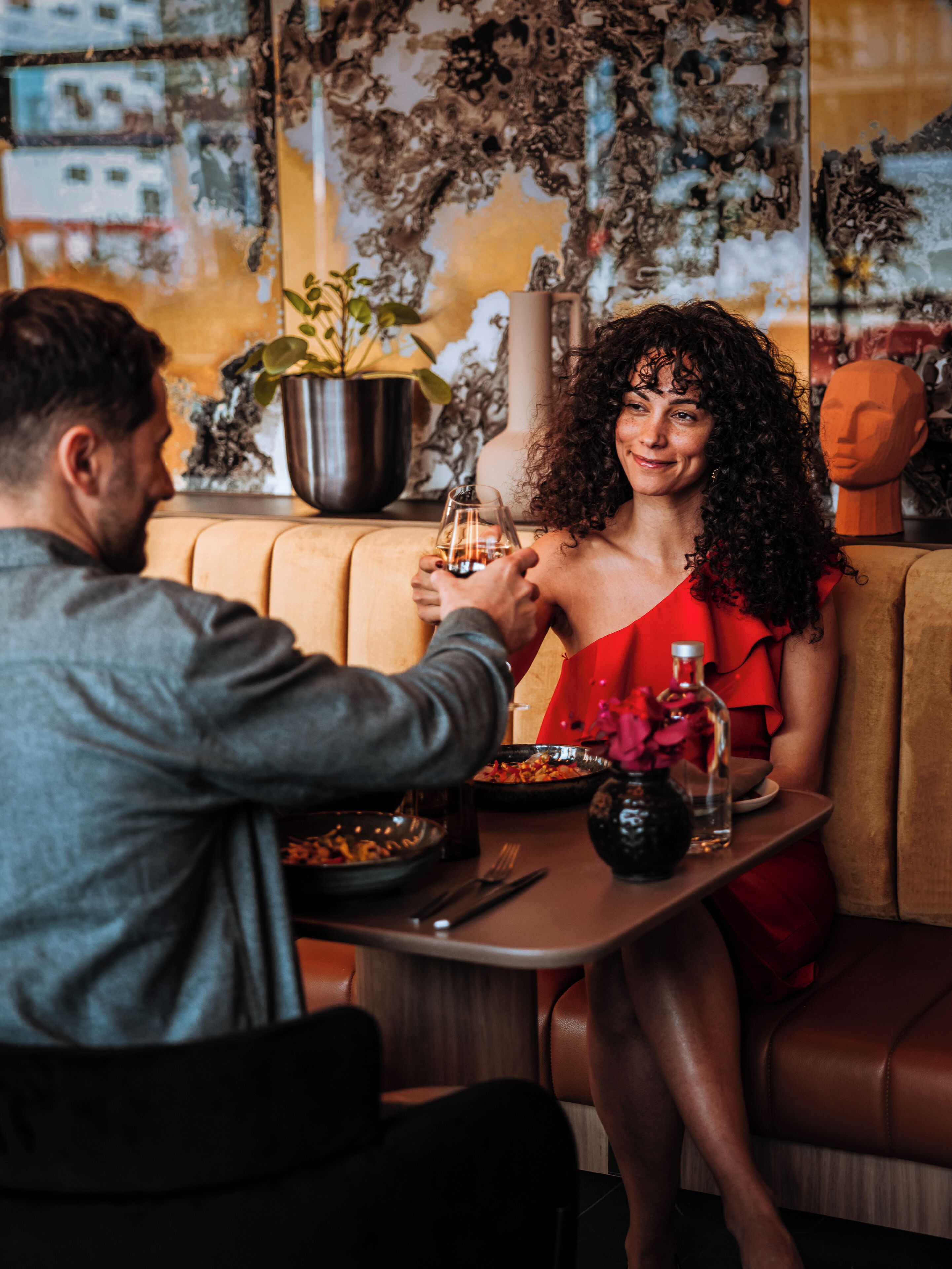 A couple seated at a table with a bottle of wine, a bowl of food, and a vase of flowers.