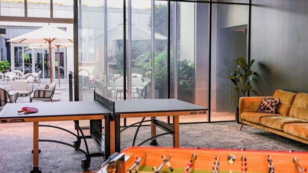 Una mesa de fútbol de mesa, un sofá y una mesa de ping pong en un espacio interior moderno con ventanas amplias.