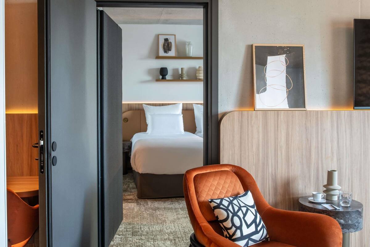 Een hotelkamer met een bed, een stoel met een gestreept kussen, een klein tafeltje met een kopje en vaas, en kunst aan de muur.