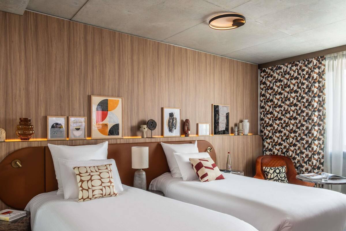 Een hotelkamer met twee bedden, een houten hoofdbord, een klein tafeltje met een lamp en kunst aan de muur.