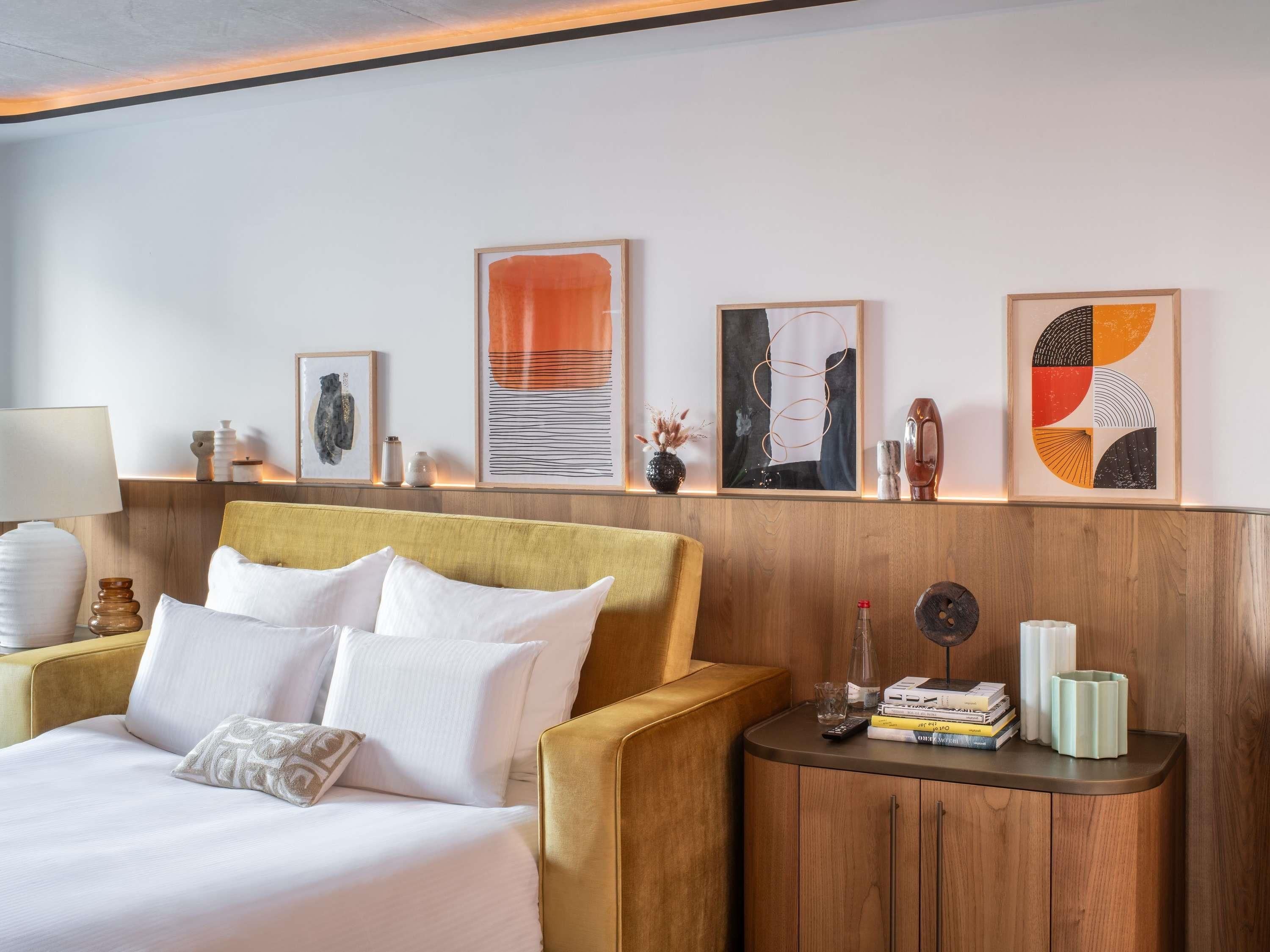 A hotel room with a bed, a sofa, a nightstand with books and a remote control, and artwork on the wall.