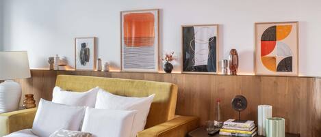 A hotel room with a bed, a sofa, a nightstand with books and a remote control, and artwork on the wall.