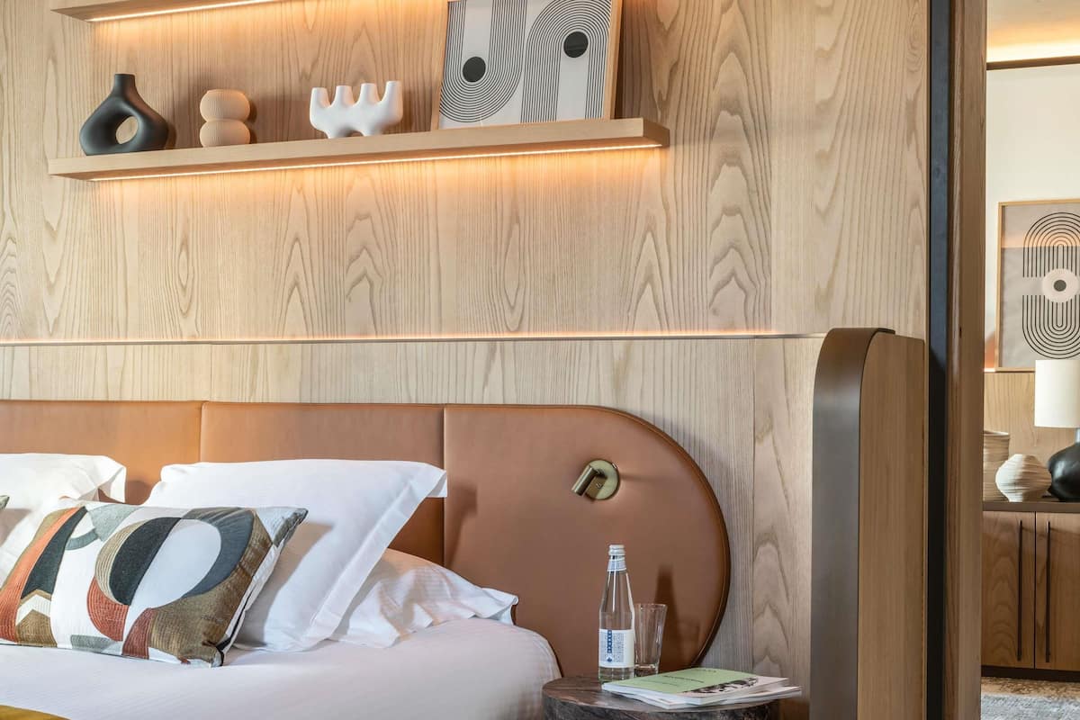 A hotel room with a bed, a nightstand, a wooden shelf with decorative items, and a wall-mounted artwork.