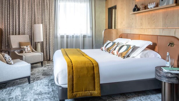 A modern hotel room with a large bed, a seating area with a chair and a table, and a shelf with decorative items.